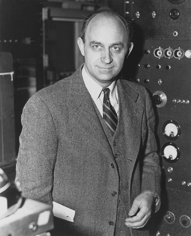 Black-and-white portrait of Enrico Fermi in a tweed jacket, standing in front of a laboratory control panel